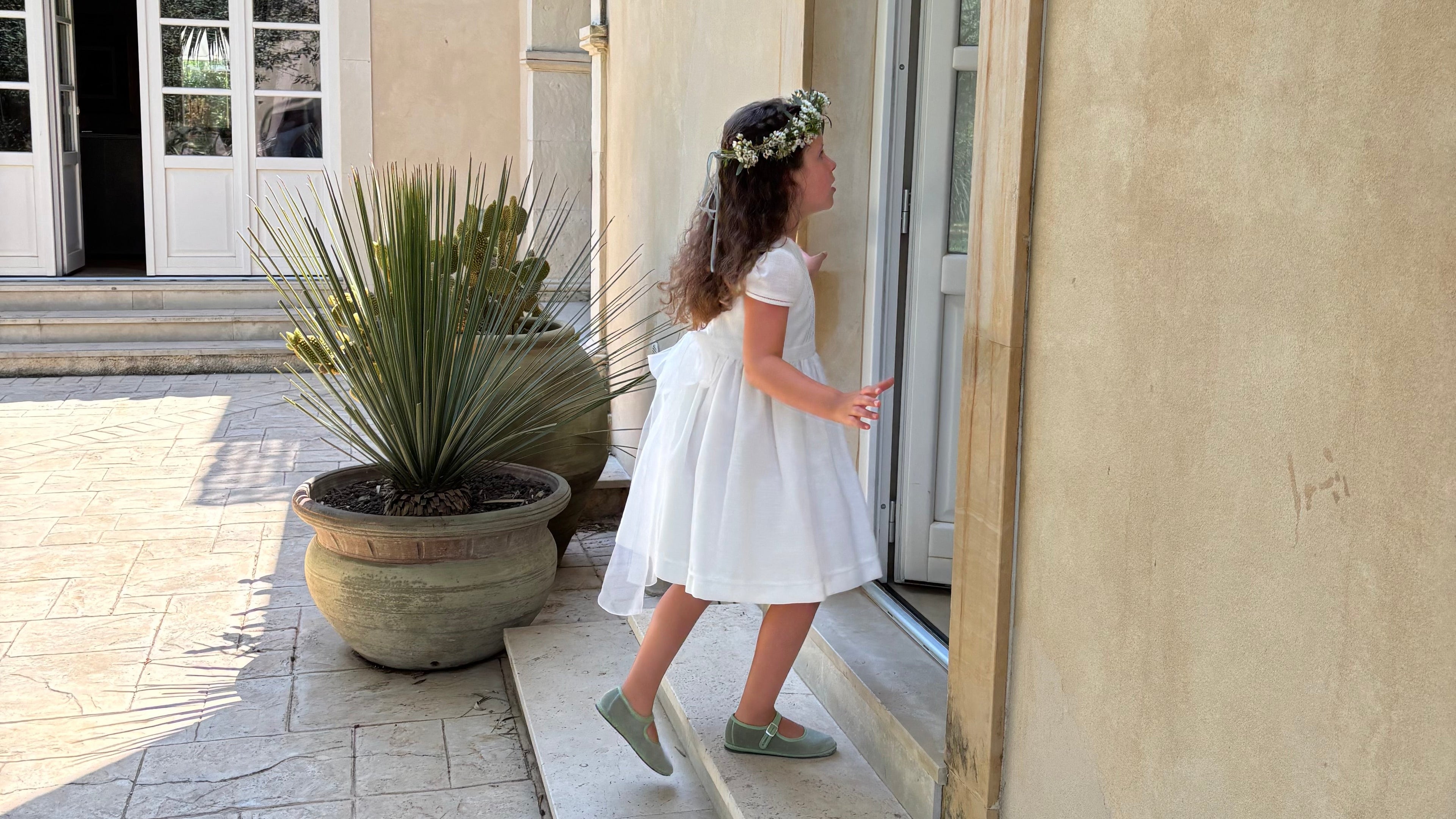 Young girl in a white dress standing on a stone path outside a building with plants and a clear sky.